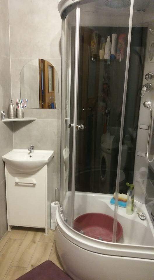 Ukończona łazienka z narożną kabiną prysznicową z hydromasażem, umywalką z szafką i półką z akcesoriami, w tle widoczna pralka.