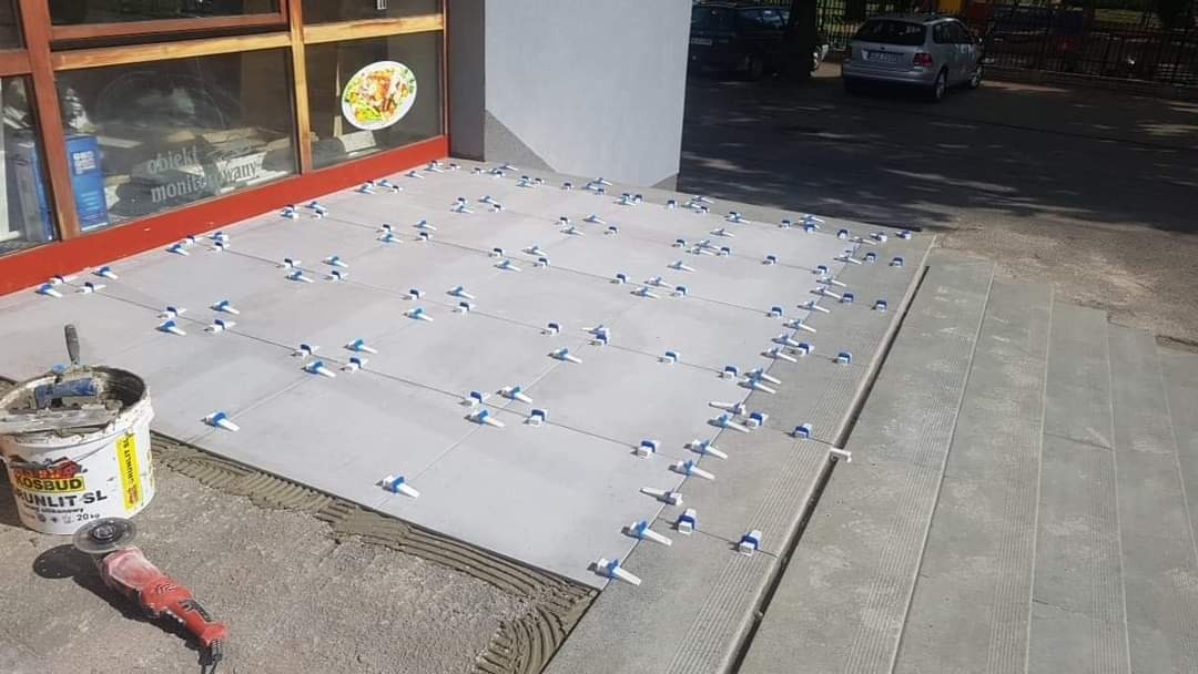 Układanie jasnoszarych płytek na zewnątrz budynku z użyciem systemu poziomowania; widoczne narzędzia, wiadro z klejem Kosbud Runlit SL oraz szlifierka kątowa; w tle okno wystawowe i zaparkowany...