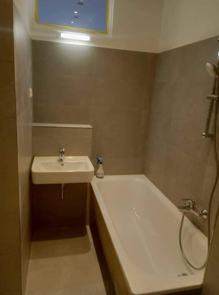 Nowoczesna łazienka z beżowymi płytkami, białą wanną i umywalką na metalowym stelażu, uzupełniona o okno dachowe z żółtą ramą i butelkę z rozpylaczem na wannie.