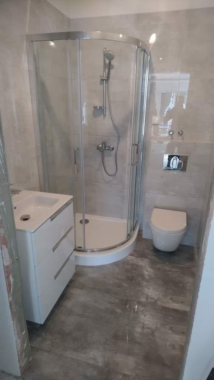 Nowoczesna łazienka z kabiną prysznicową z zaokrąglonymi drzwiami, umywalką z szafką z trzema szufladami, podwieszaną toaletą i szarymi płytkami imitującymi beton na ścianach i podłodze.