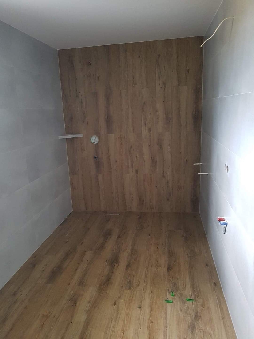 Wnętrze łazienki w trakcie wykańczania: drewnopodobne panele na podłodze i ścianie, białe płytki na ścianach, widoczne wyprowadzenia instalacji wodnej i elektrycznej, niewielka półka. Na podłodze...