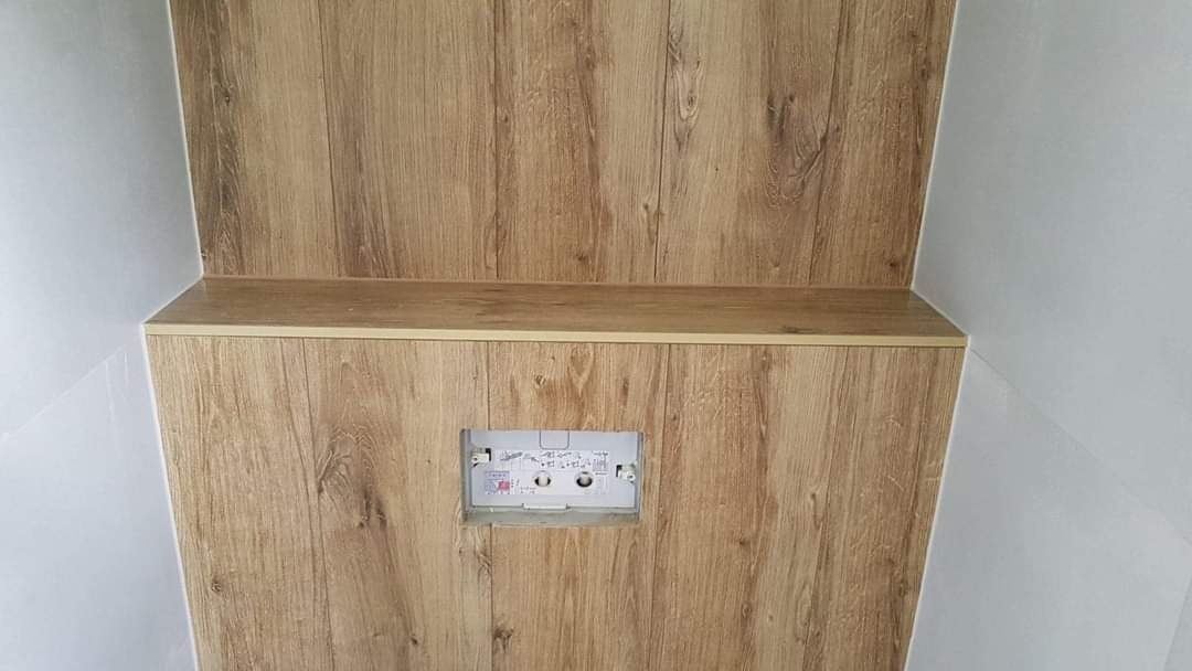 Wykończenie ściany i zabudowy stelaża WC w łazience, panele imitujące drewno, widoczne przyłącza hydrauliczne i elektryczne w otworze montażowym.
