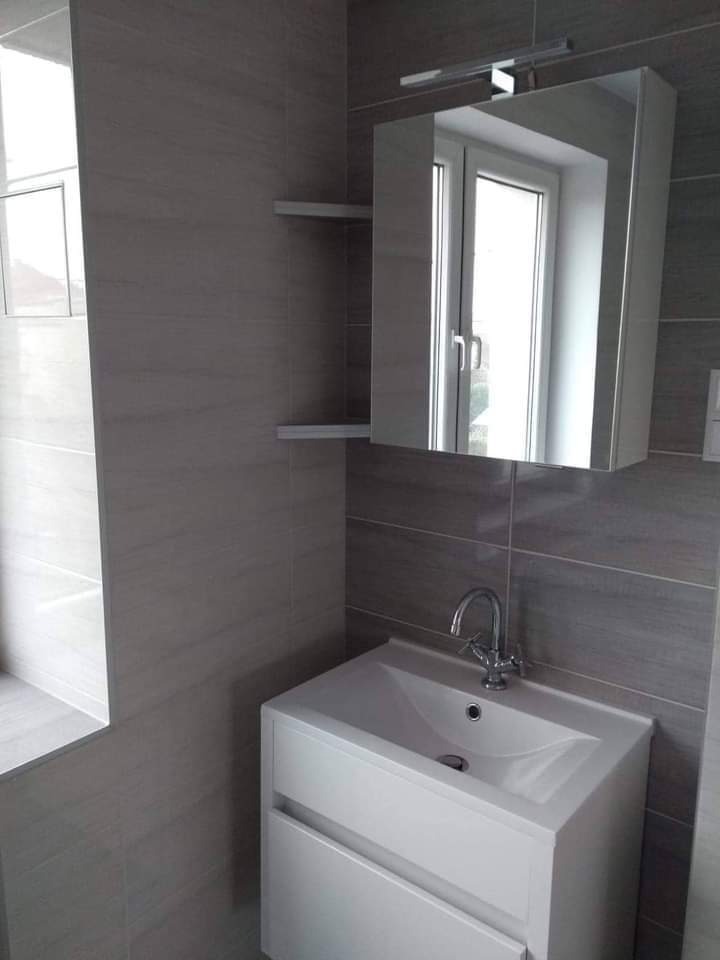 Nowoczesna łazienka z szarymi płytkami imitującymi drewno, białą szafką z umywalką i lustrem z oświetleniem LED.