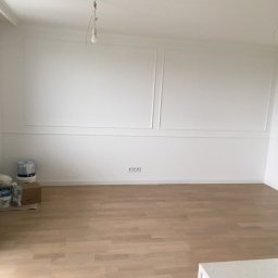 Comfort tawn - Pomalowane na biało wnętrze pokoju z drewnianą podłogą, ozdobnymi panelami ściennymi i wiszącą żarówką bez klosza. W rogu widoczne puszki z farbą i karton.