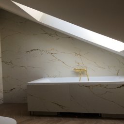 Comfort tawn - Nowoczesna łazienka na poddaszu z wanną obudowaną płytami imitującymi marmur z złotą armaturą, oświetlona przez okno dachowe.