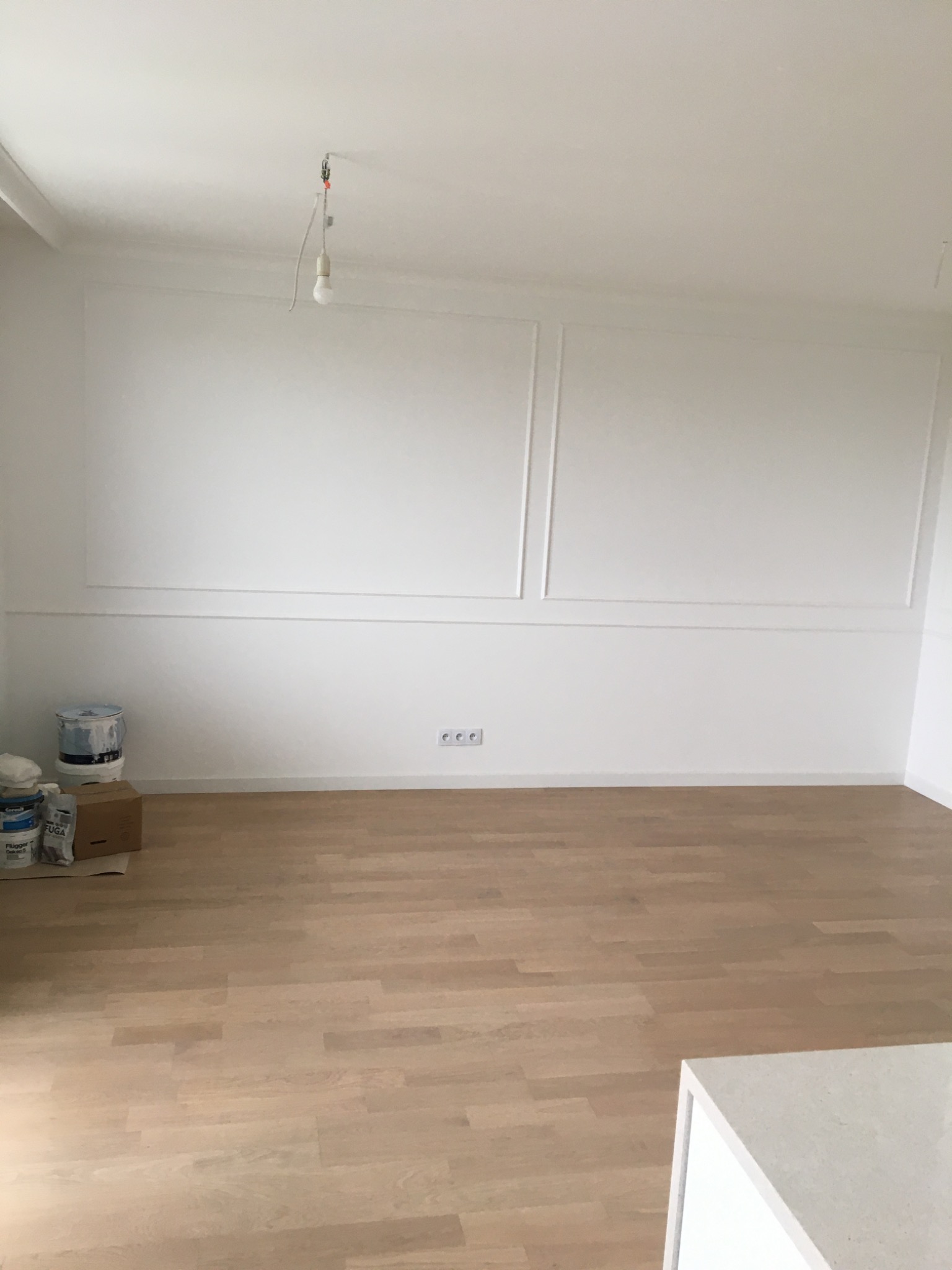 Pomalowane na biało wnętrze pokoju z drewnianą podłogą, ozdobnymi panelami ściennymi i wiszącą żarówką bez klosza. W rogu widoczne puszki z farbą i karton.