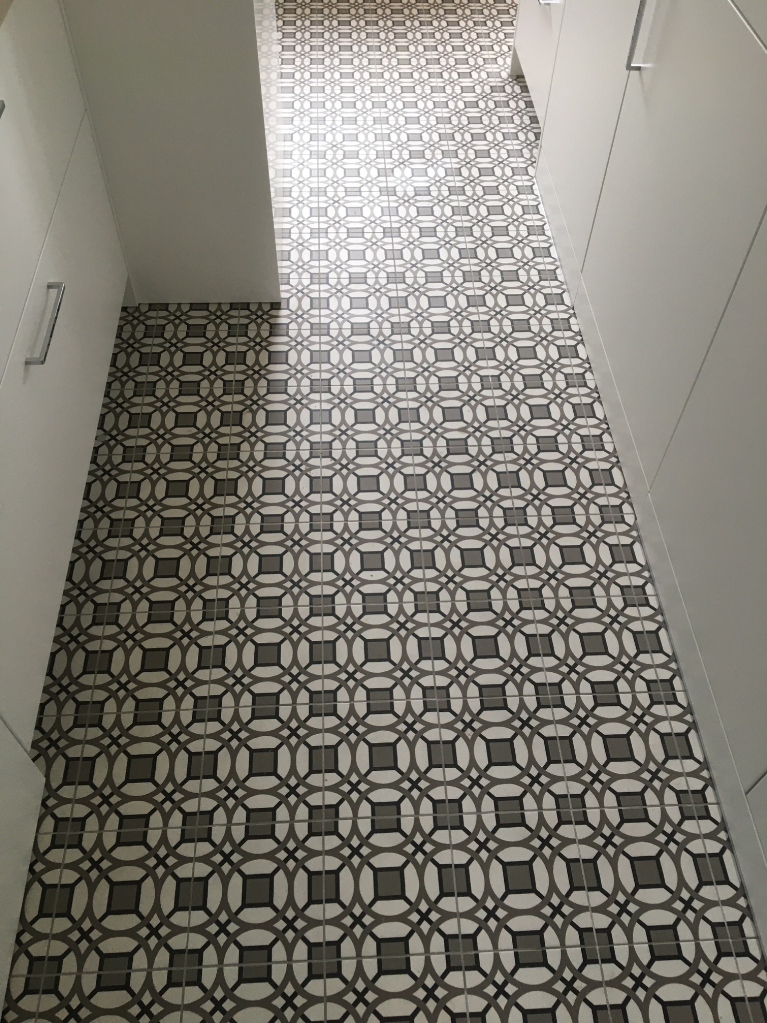 Nowoczesna podłoga w łazience wyłożona płytkami w geometryczny wzór, szare i białe płytki ułożone w kwadraty i okręgi, widoczne fragmenty białych szafek.