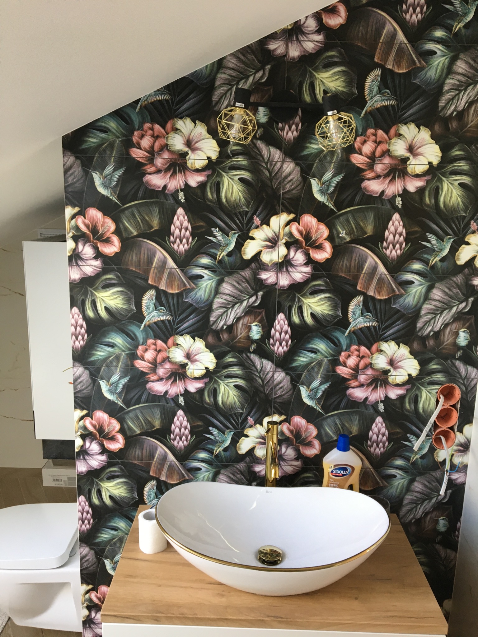 Umywalka nablatowa z baterią w kolorze złotym na blacie z drewnopodobnego materiału, tło stanowi ściana wyłożona płytkami z motywem tropikalnych kwiatów i kolibrów, widoczne elementy instalacji...