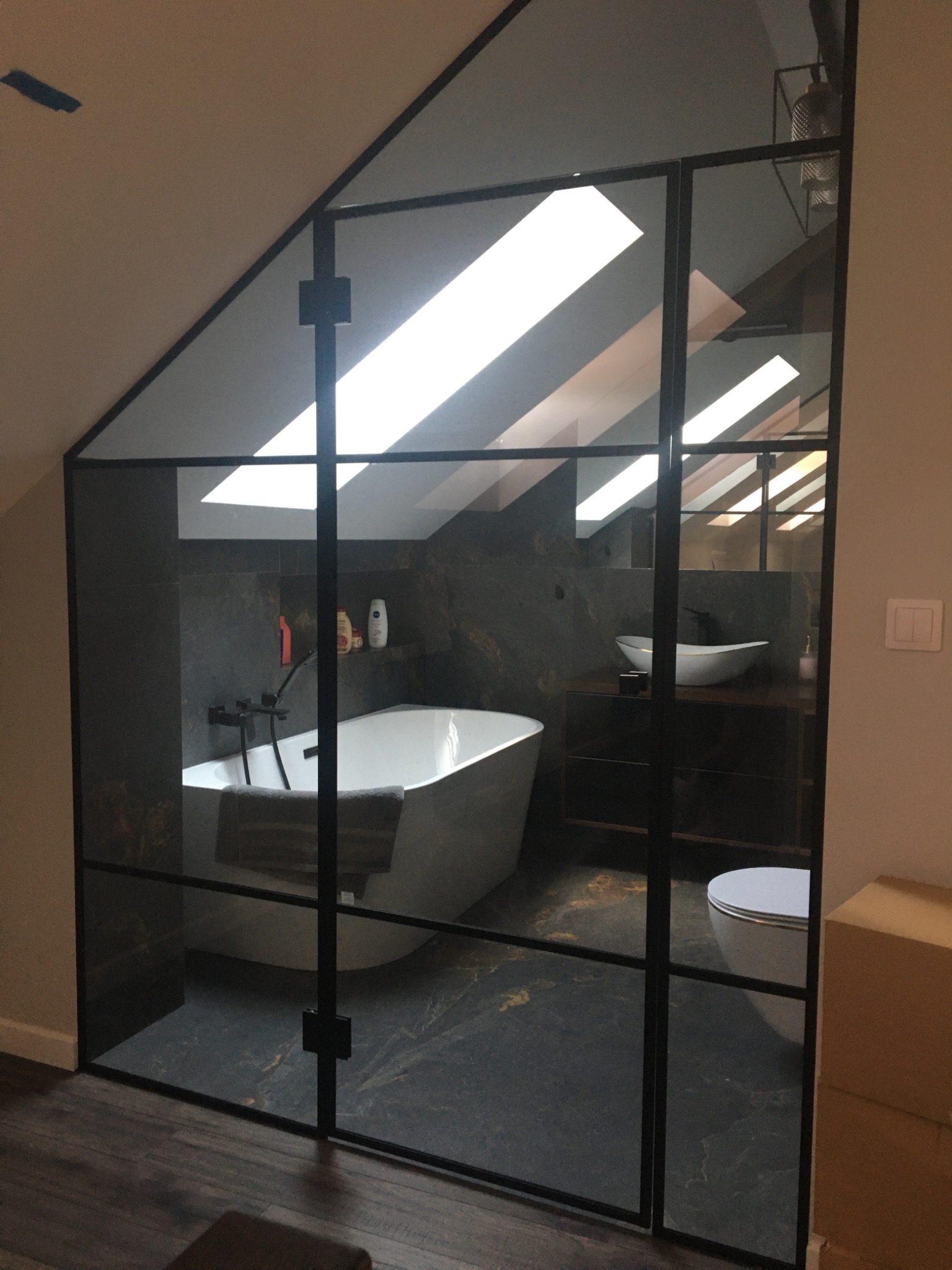 Nowoczesna łazienka na poddaszu z kabiną prysznicową typu walk-in z czarnymi profilami, białą wanną wolnostojącą i umywalką nablatową na ciemnej szafce.