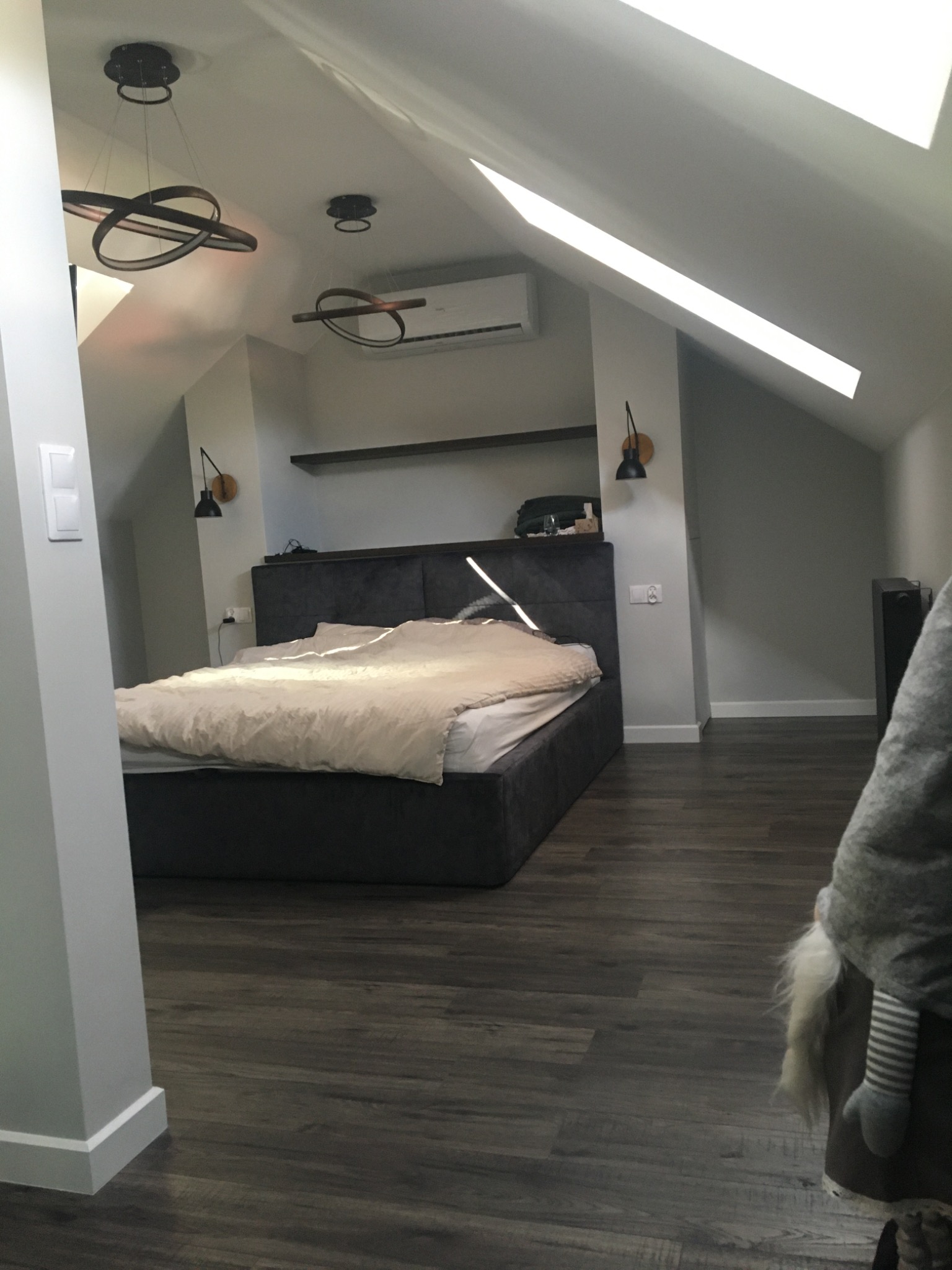 Sypialnia na poddaszu z ciemną drewnianą podłogą, szarą tapicerowaną ramą łóżka i skośnym sufitem z oknem dachowym, oświetlona nowoczesnymi lampami wiszącymi