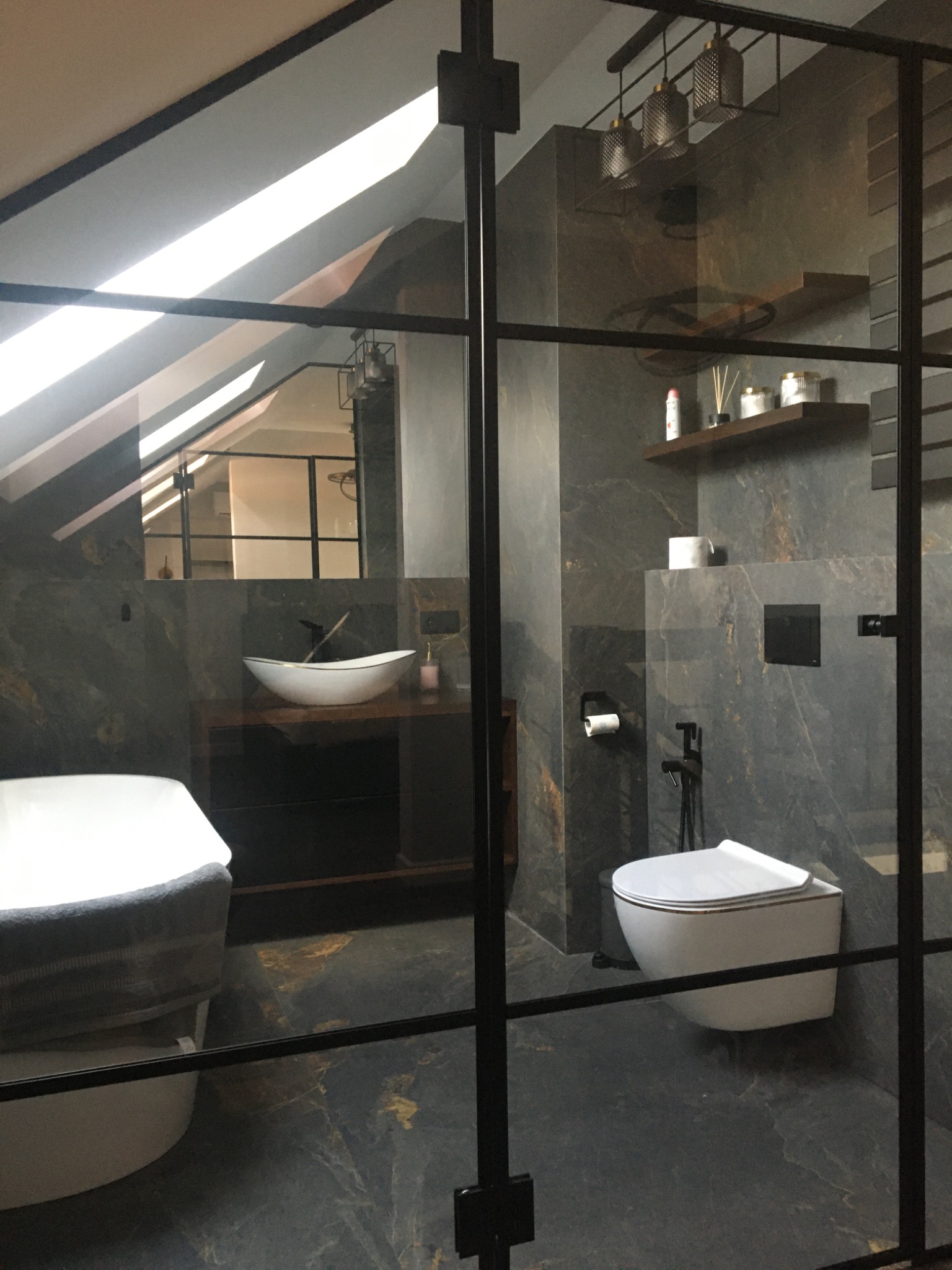 Nowoczesna łazienka w odcieniach szarości i czerni, z kabiną prysznicową typu walk-in z czarnymi szprosami, wolnostojącą wanną z szarym ręcznikiem i umywalką nablatową na ciemnej szafce, oświetlona...