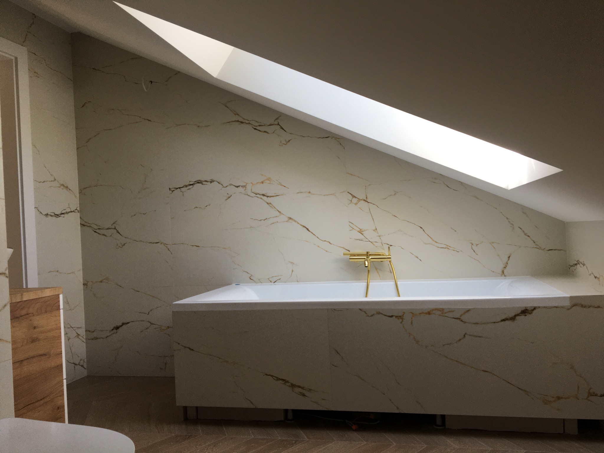 Nowoczesna łazienka na poddaszu z wanną obudowaną płytami imitującymi marmur z złotą armaturą, oświetlona przez okno dachowe.