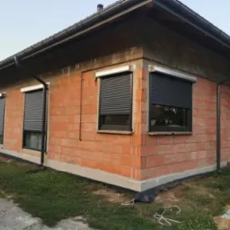 Niedokończony dom z cegły z zamontowanymi grafitowymi roletami zewnętrznymi na oknach, widoczne elementy instalacji i rynny.