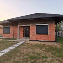 JBM Jakub Bizoń - Dom w stanie surowym w Sędziszowie Małopolskim z zamontowanymi, zewnętrznymi, czarnymi roletami na oknach, widok z przodu na trawnik i ścieżkę z płyt.