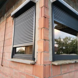 JBM Jakub Bizoń - Dwa okna z zamontowanymi, częściowo opuszczonymi, grafitowymi roletami zewnętrznymi, widoczne na tle surowej, ceglanej elewacji budynku w trakcie budowy.