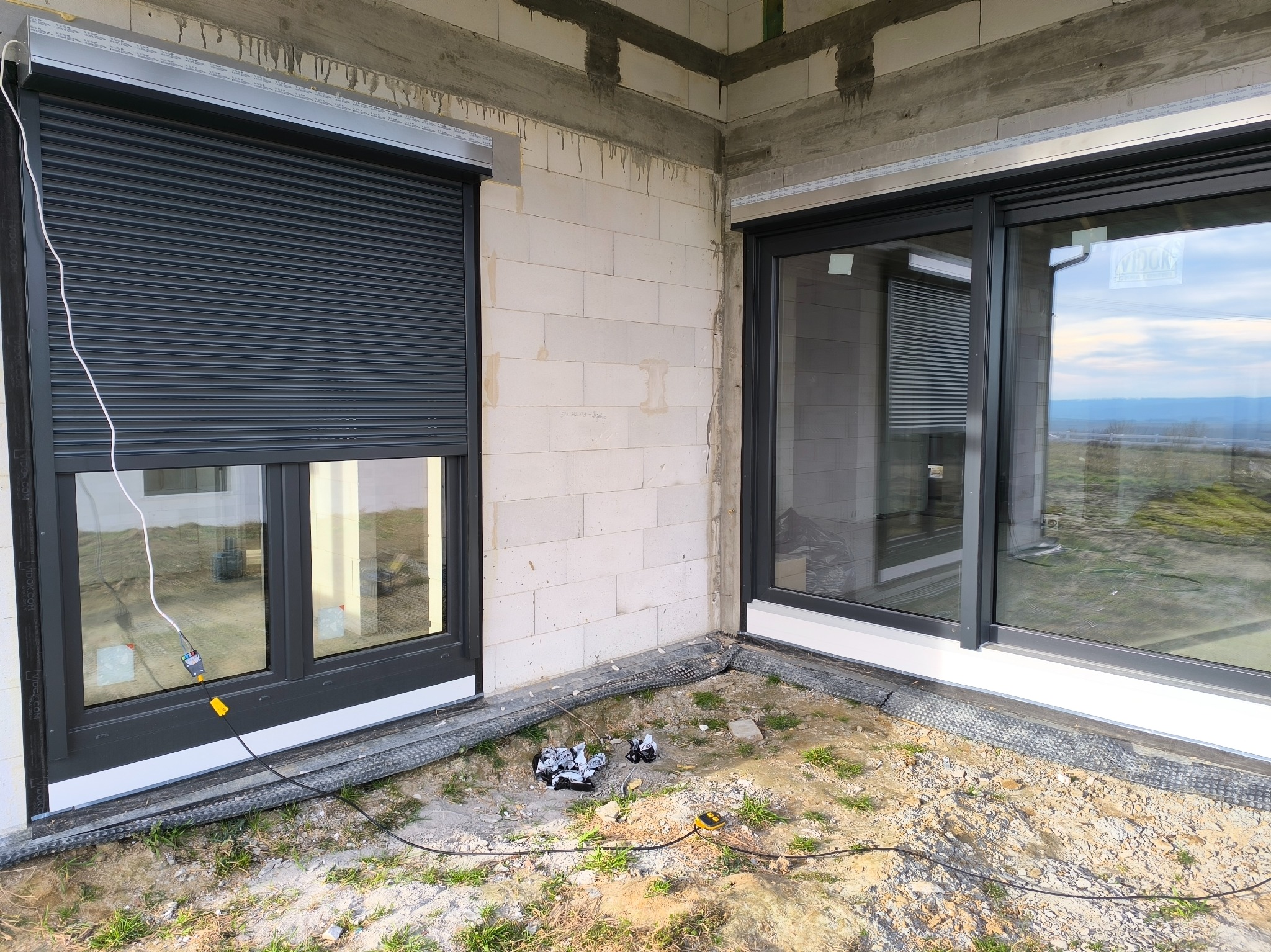 Montaż zewnętrznych rolet na surowej elewacji budynku w trakcie budowy, widoczne narzędzia i przewody elektryczne.