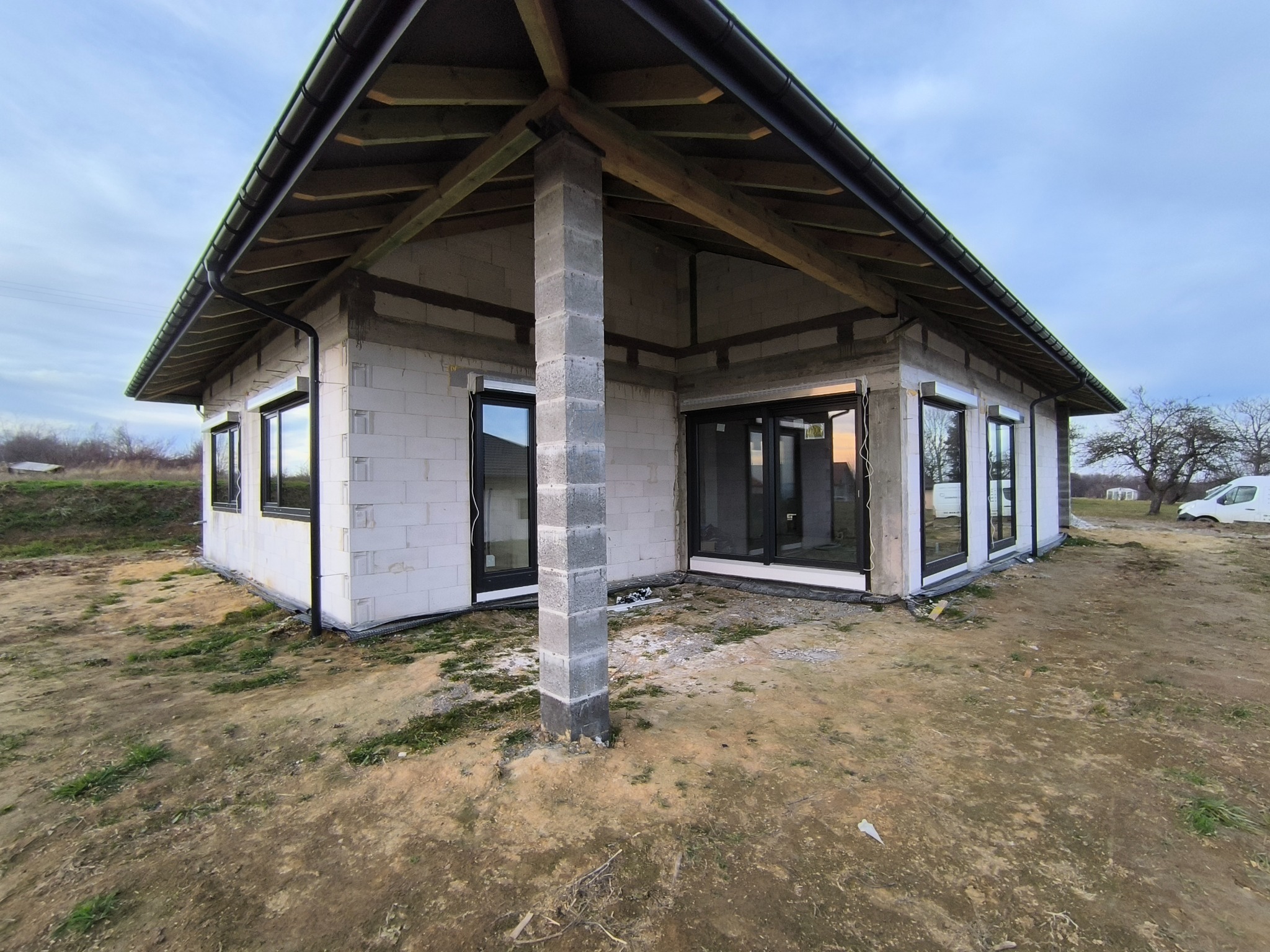 Surowy stan domu jednorodzinnego z białego bloczka, widoczne okna z zamontowanymi zewnętrznymi osłonami okiennymi w kolorze czarnym, teren wokół domu niezagospodarowany.