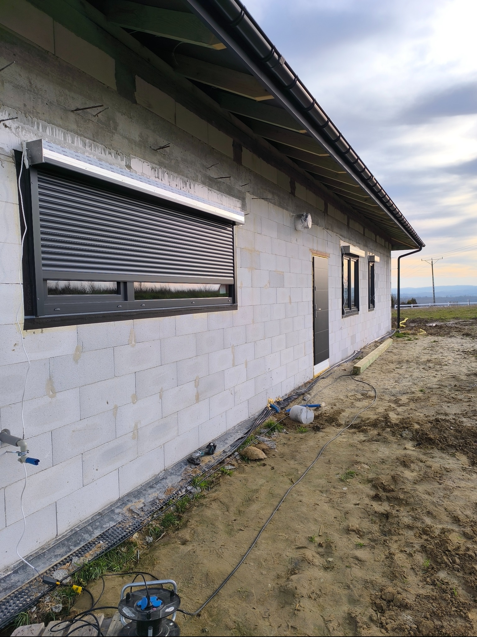 Szara roleta zewnętrzna częściowo opuszczona na oknie domu w trakcie budowy, widoczne bloczki betonowe i przygotowany teren.