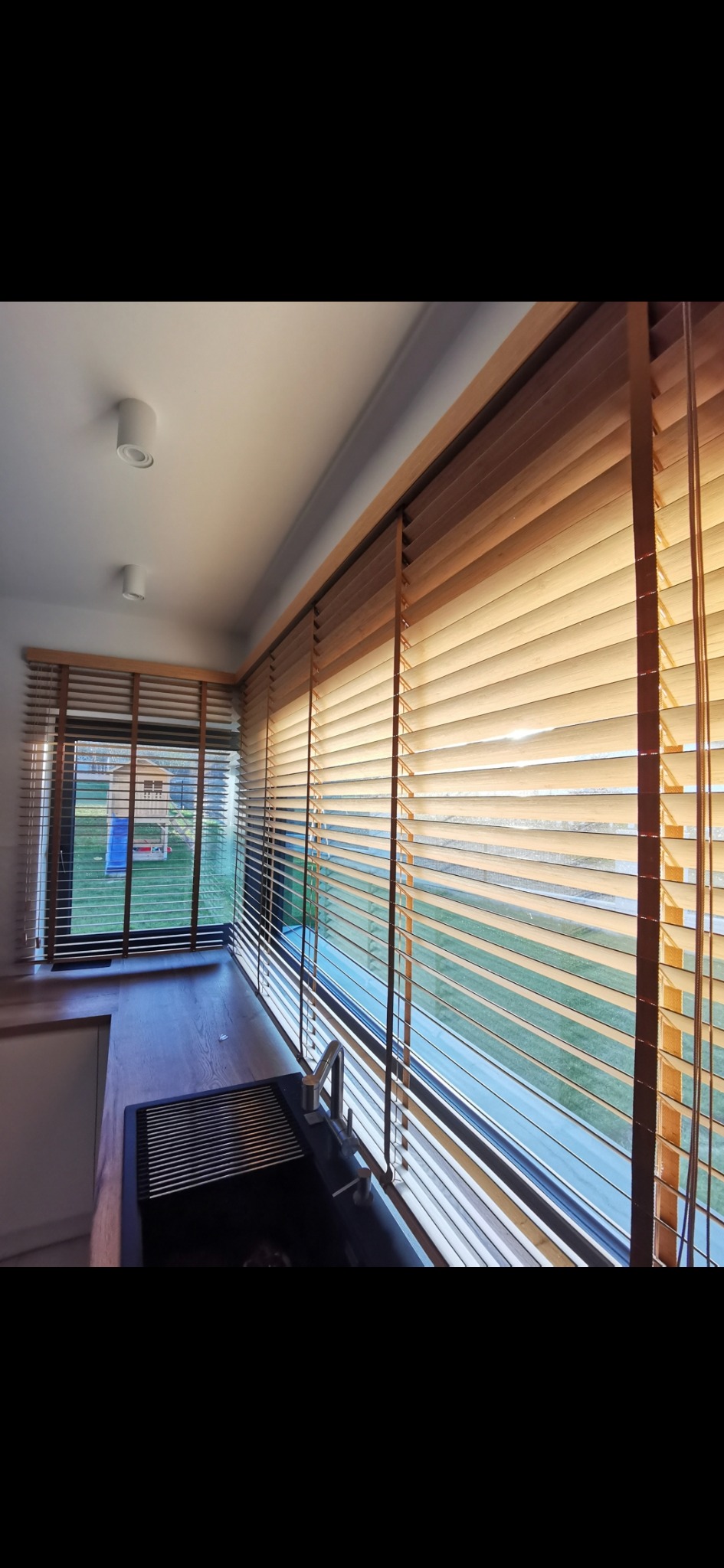 Drewniane żaluzje poziome w kuchni, widok na ogród przez okno, czarny zlew z ociekaczem.