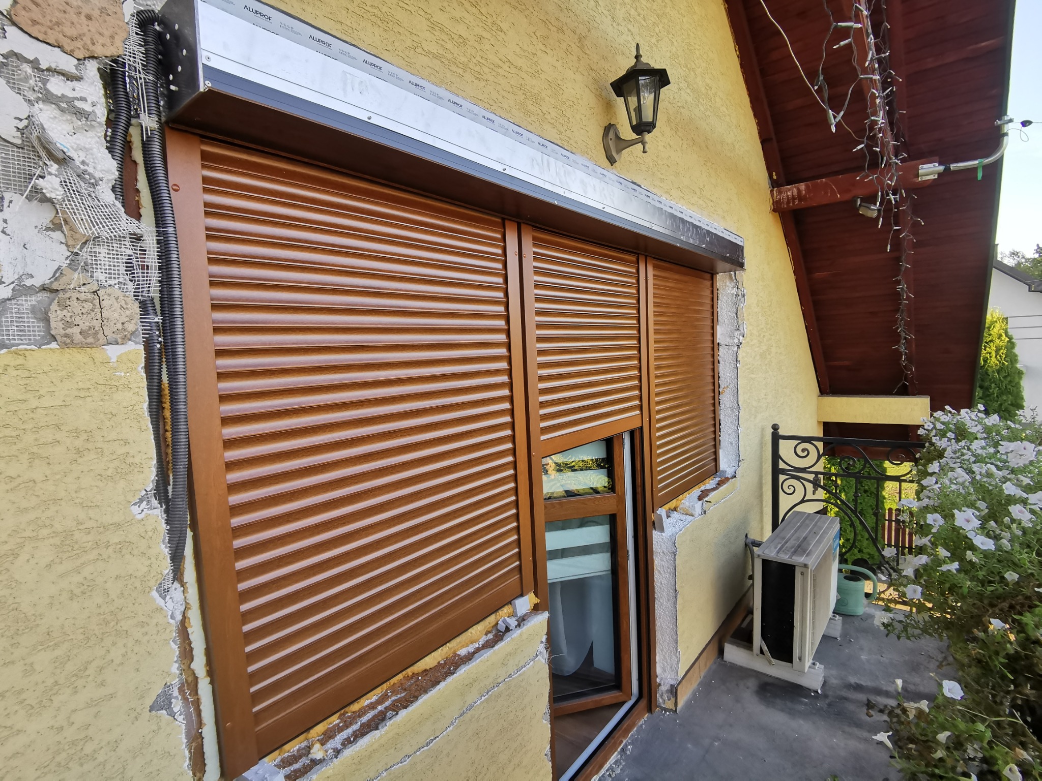 Brązowe rolety zewnętrzne zamontowane na oknie domu z żółtą elewacją, widoczne ślady po montażu na ścianie, oświetlenie zewnętrzne i klimatyzator na zewnątrz.