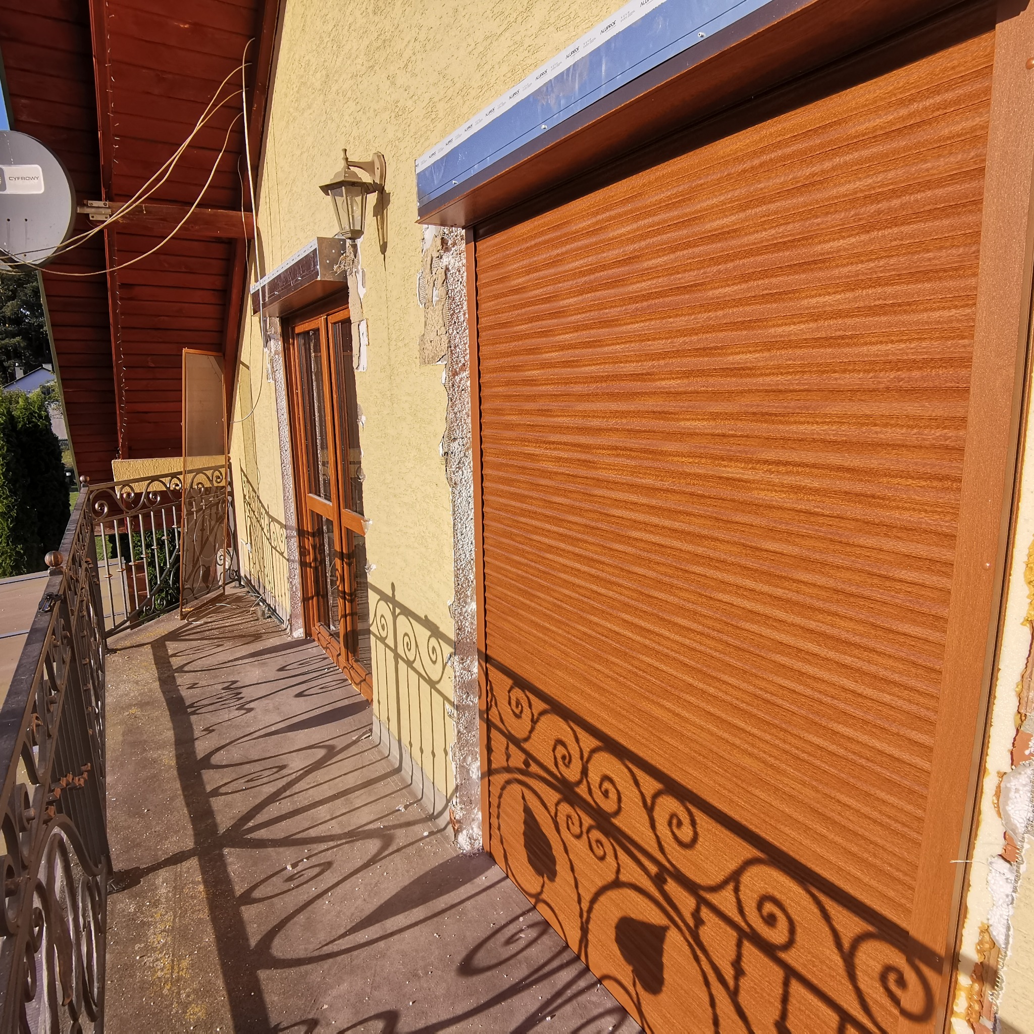 Zamknięta brązowa roleta zewnętrzna zamontowana na budynku z balkonem, widoczne niedokończone prace wykończeniowe wokół okna i rolety.