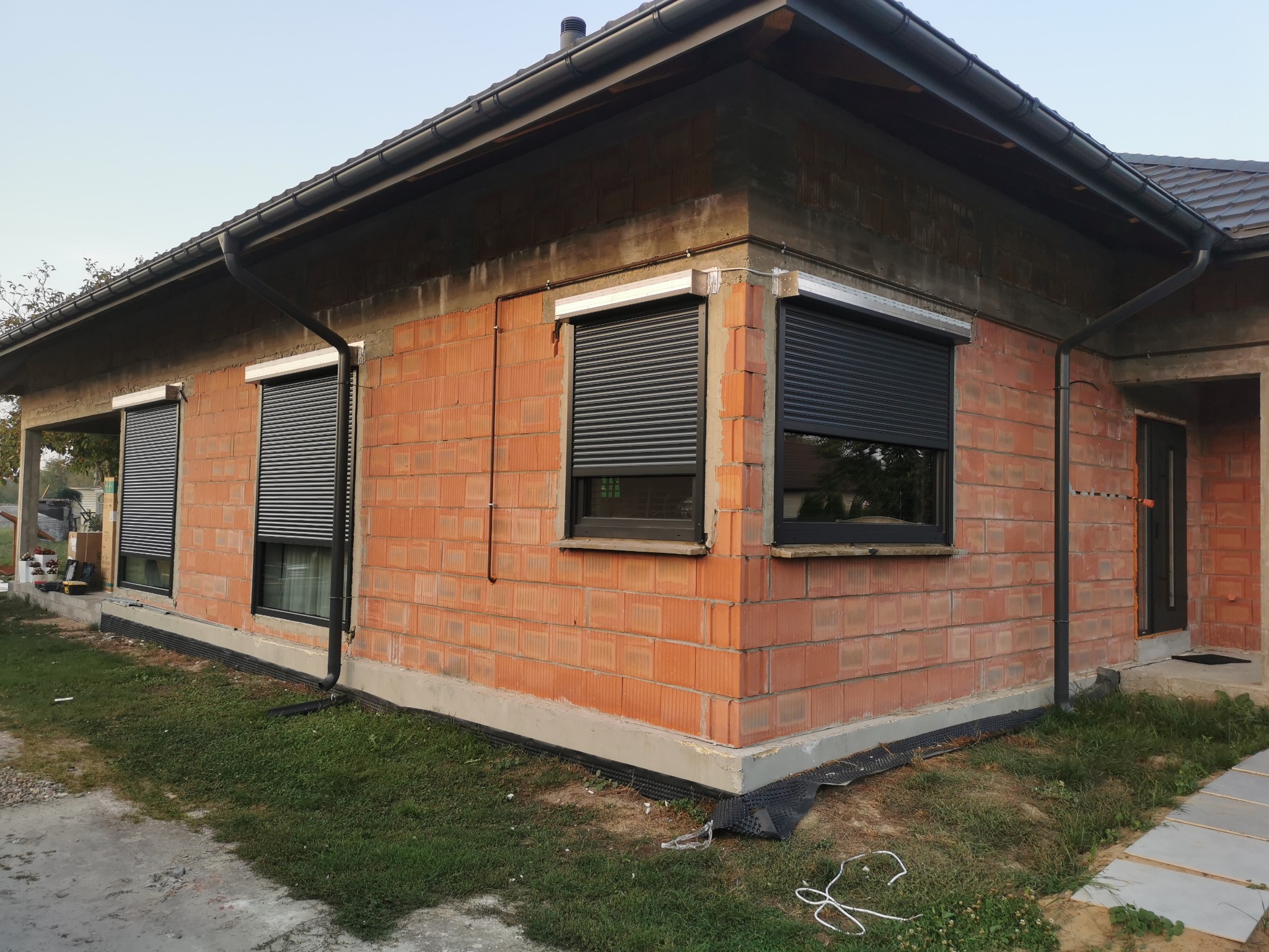 Niedokończony dom z cegły z zamontowanymi grafitowymi roletami zewnętrznymi na oknach, widoczne elementy instalacji i rynny.