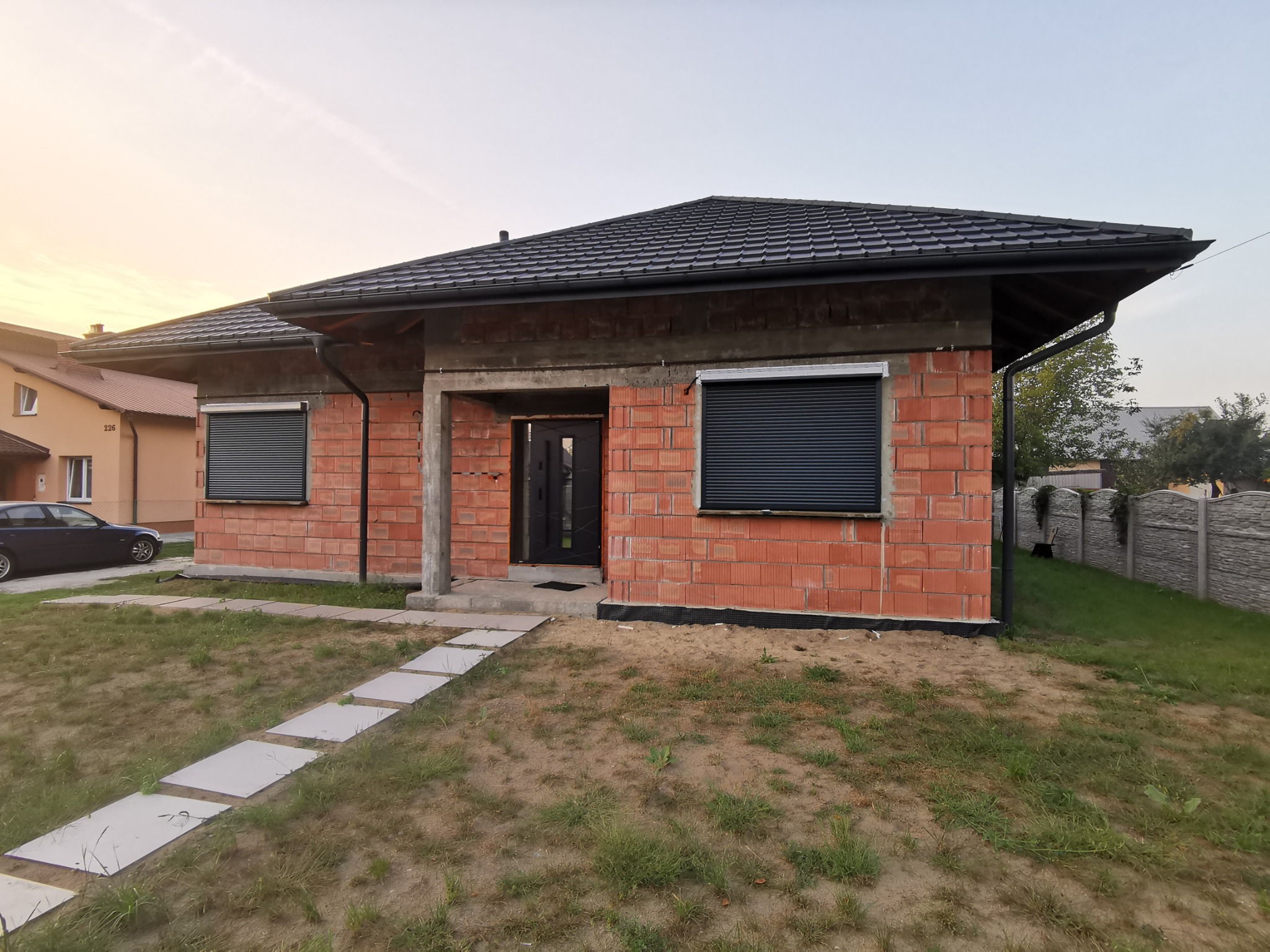 Dom w stanie surowym w Sędziszowie Małopolskim z zamontowanymi, zewnętrznymi, czarnymi roletami na oknach, widok z przodu na trawnik i ścieżkę z płyt.