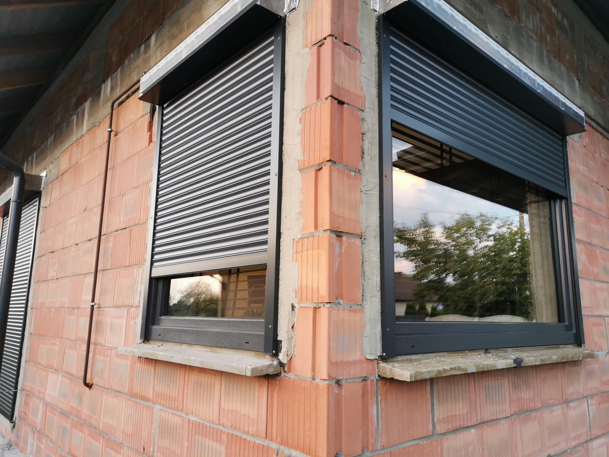 Dwa okna z zamontowanymi, częściowo opuszczonymi, grafitowymi roletami zewnętrznymi, widoczne na tle surowej, ceglanej elewacji budynku w trakcie budowy.