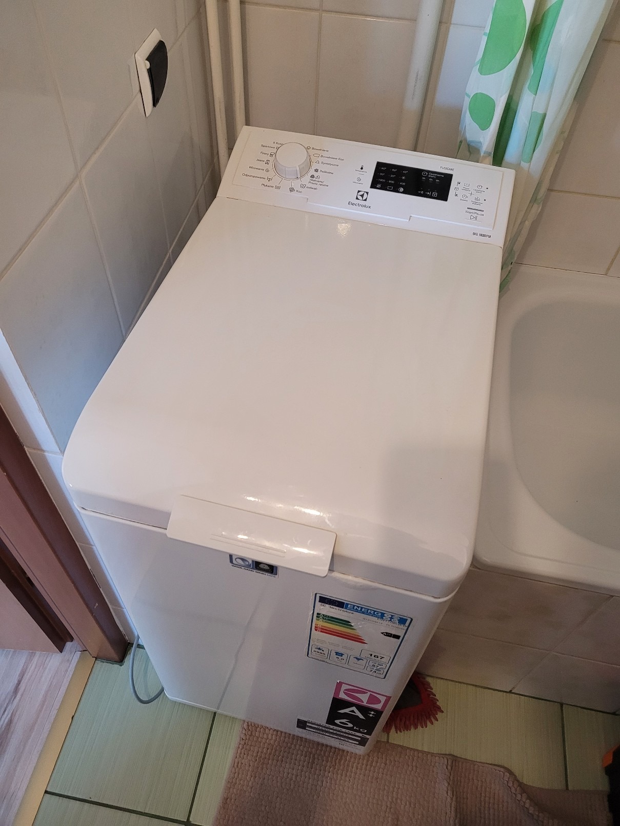 Biała pralka ładowana od góry marki Electrolux w łazience, widoczne płytki ceramiczne na ścianach i zasłonka prysznicowa w tle. Panel sterowania z pokrętłem i wyświetlaczem cyfrowym.