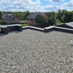 Płaski dach pokryty drobnymi kamieniami, widoczne kominki wentylacyjne i obróbki blacharskie, w tle zabudowa jednorodzinna i zieleń.