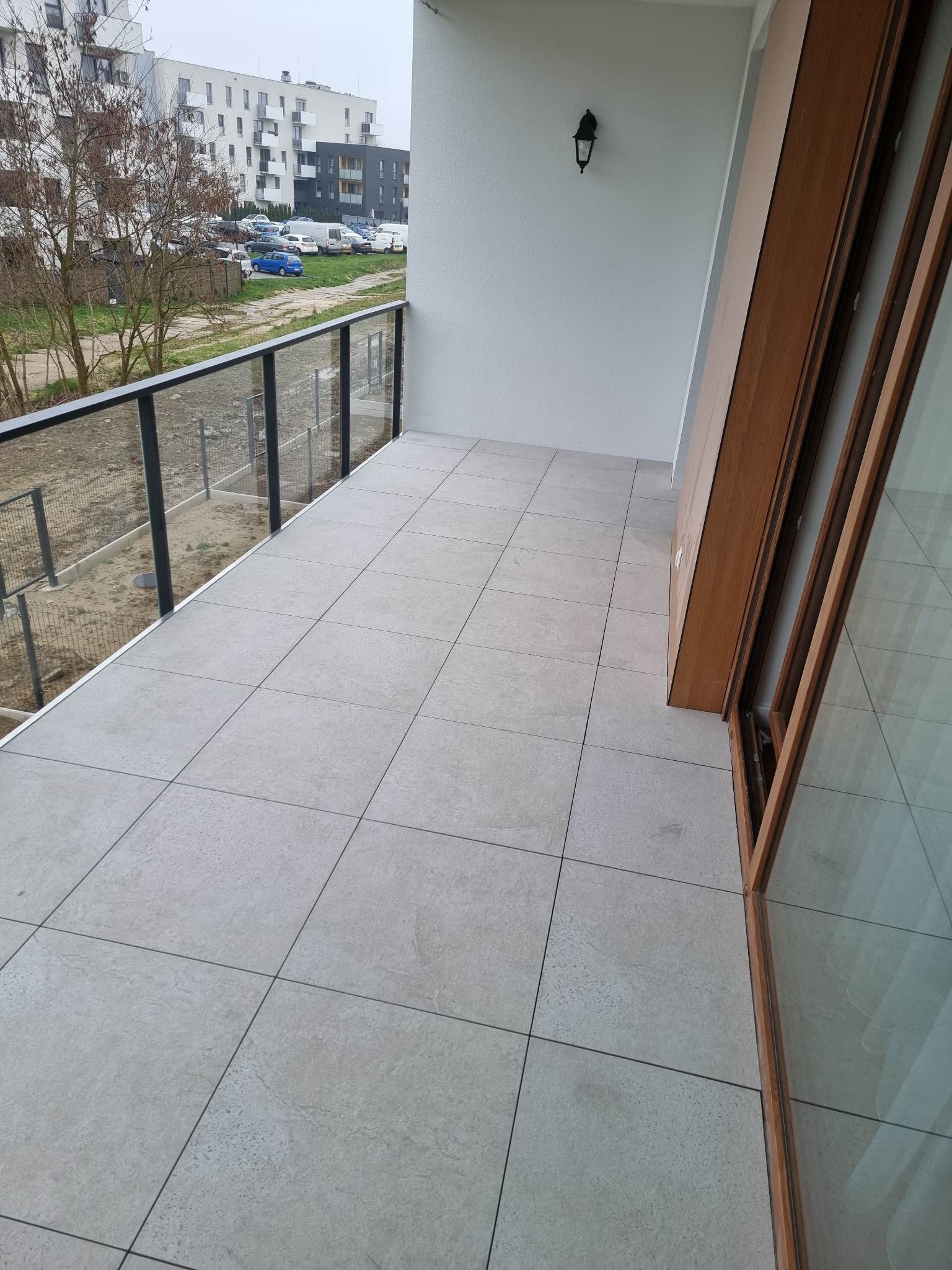 Wyłożony szarymi płytkami balkon z czarną, przeszkloną balustradą. Na ścianie lampa w stylu retro, widok na parking i budynki mieszkalne.