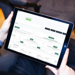 Osoba trzyma tablet prezentujący interfejs aplikacji do kalkulacji zapotrzebowania energetycznego budynku, z danymi technicznymi, mocą grzewczą i rocznym zużyciem energii.