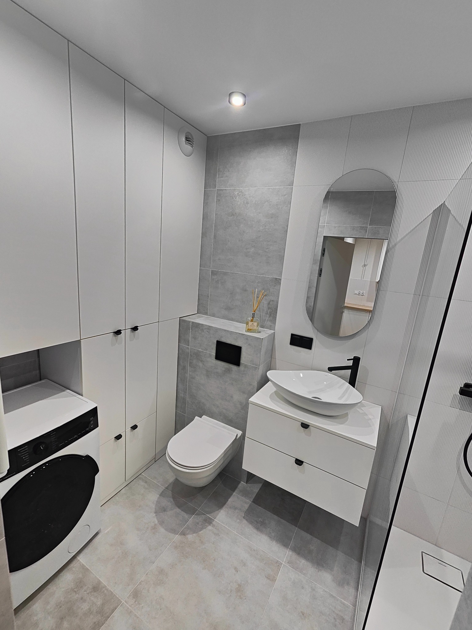 Nowoczesna łazienka w odcieniach szarości i bieli z pralką, umywalką nablatową, toaletą podwieszaną i dużym lustrem o zaokrąglonym kształcie.