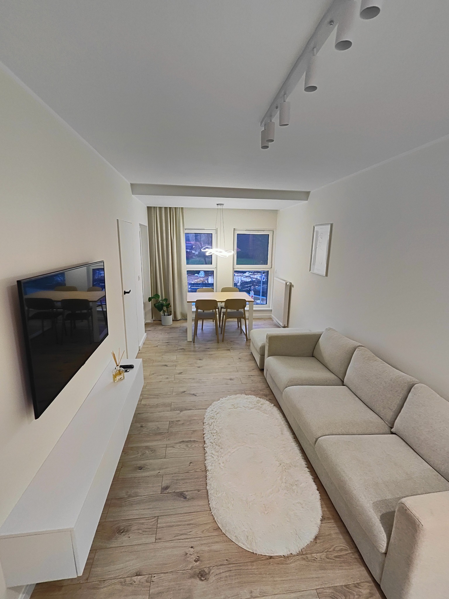 Jasne, przestronne wnętrze mieszkania po remoncie, z drewnopodobną podłogą, szarą sofą, białymi meblami RTV i widoczną częścią jadalni z oknem.