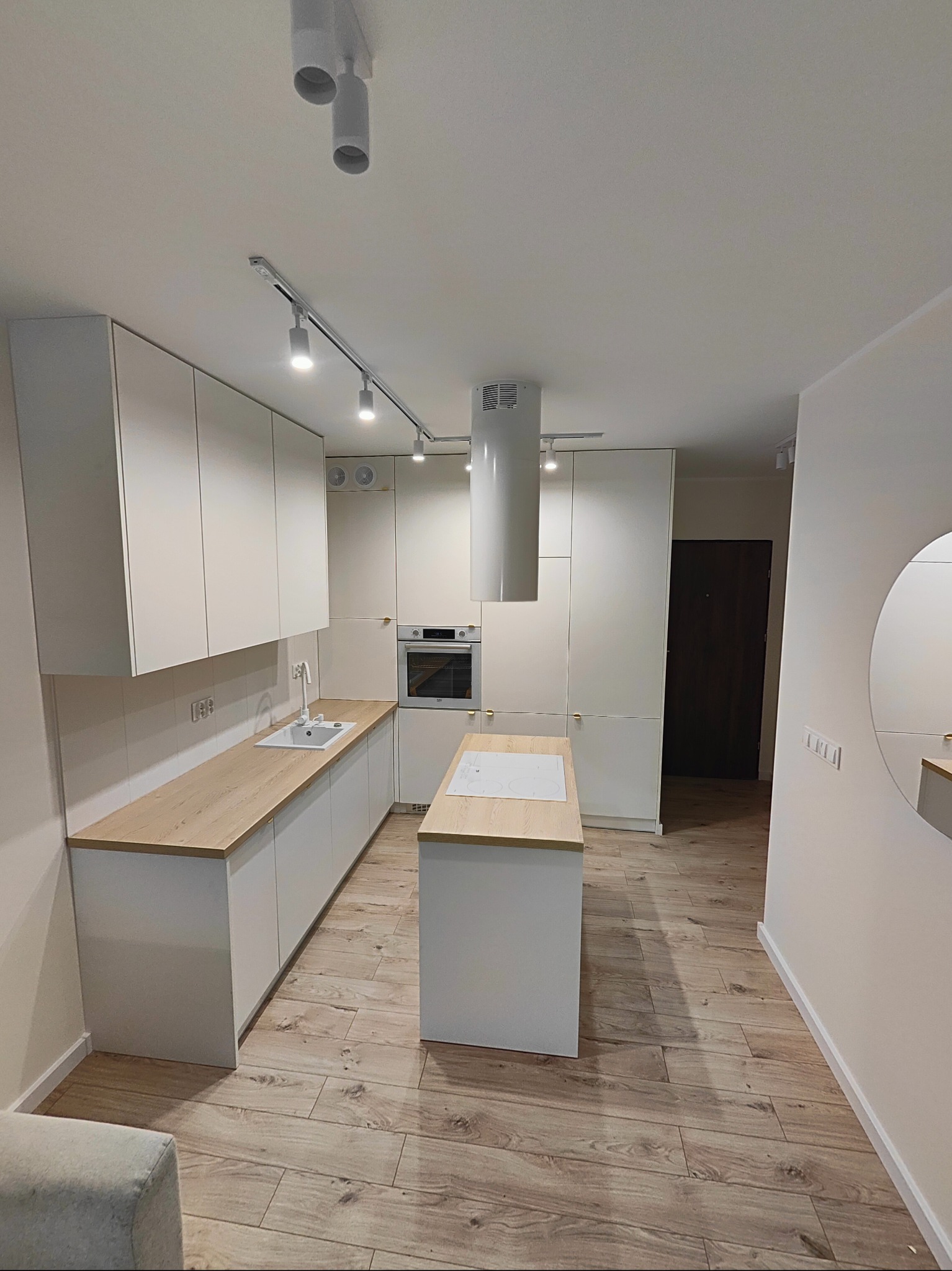 Nowoczesna, jasna kuchnia z białymi szafkami i drewnianym blatem, wyspą kuchenną i podłogą imitującą drewno, oświetlona szynoprzewodem.