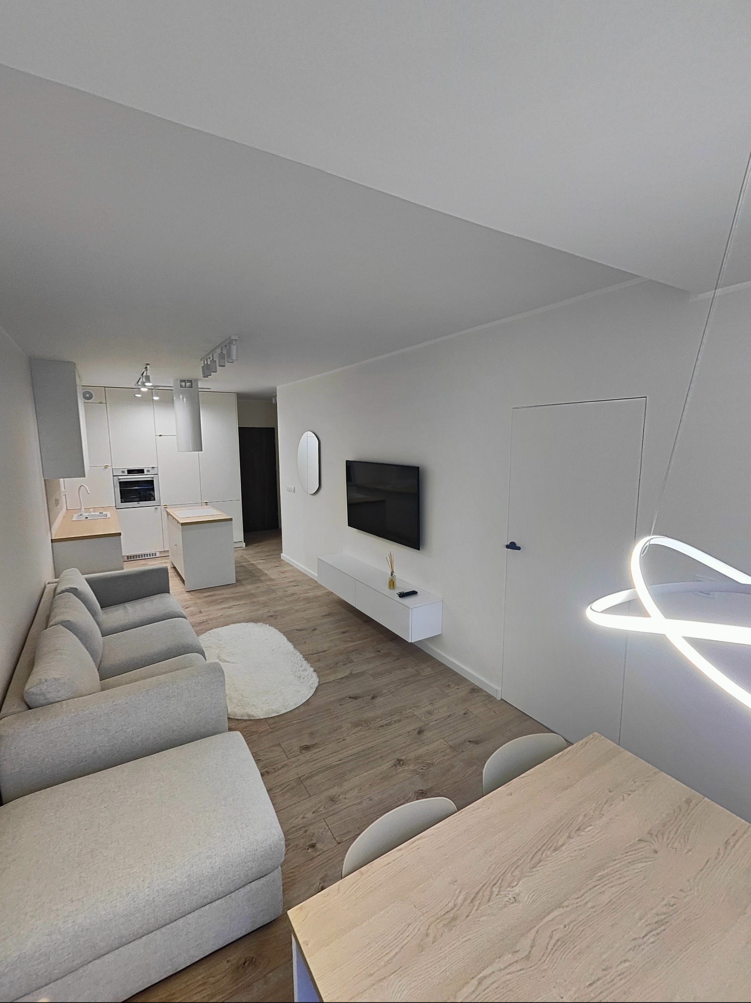 Jasne, minimalistyczne wnętrze mieszkania z drewnianą podłogą, szarą sofą, białymi meblami kuchennymi i designerską lampą nad stołem. Widoczny telewizor na ścianie i okrągły, puszysty dywan.