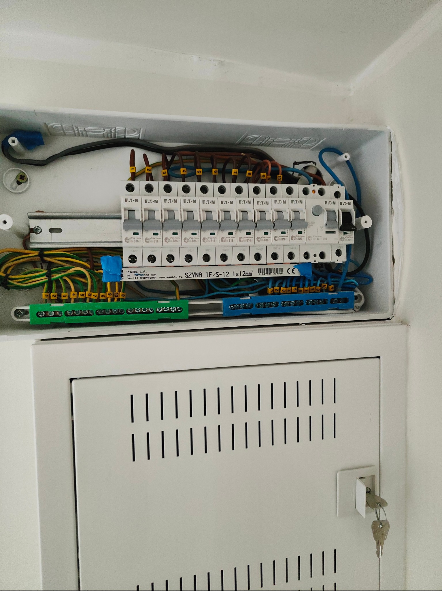 Otwarta skrzynka elektryczna z widocznymi bezpiecznikami, przewodami i złączkami, zamontowana w białej obudowie zamykanej na klucz.