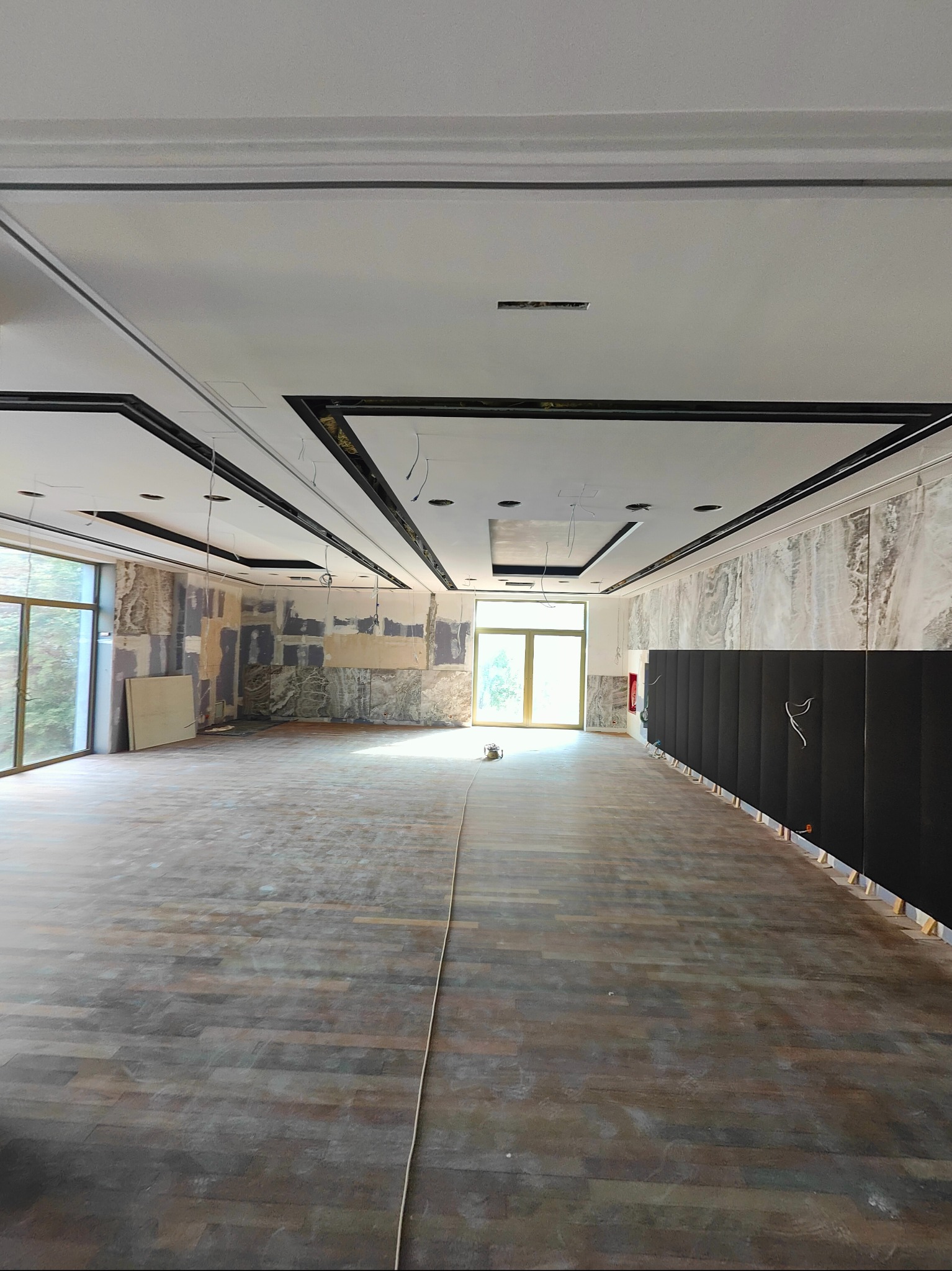 Przestronne wnętrze hotelowe w trakcie prac wykończeniowych: widoczne podwieszane sufity z czarnymi obramowaniami, surowe ściany częściowo pokryte płytami, drewnopodobna podłoga oraz czarne panele...