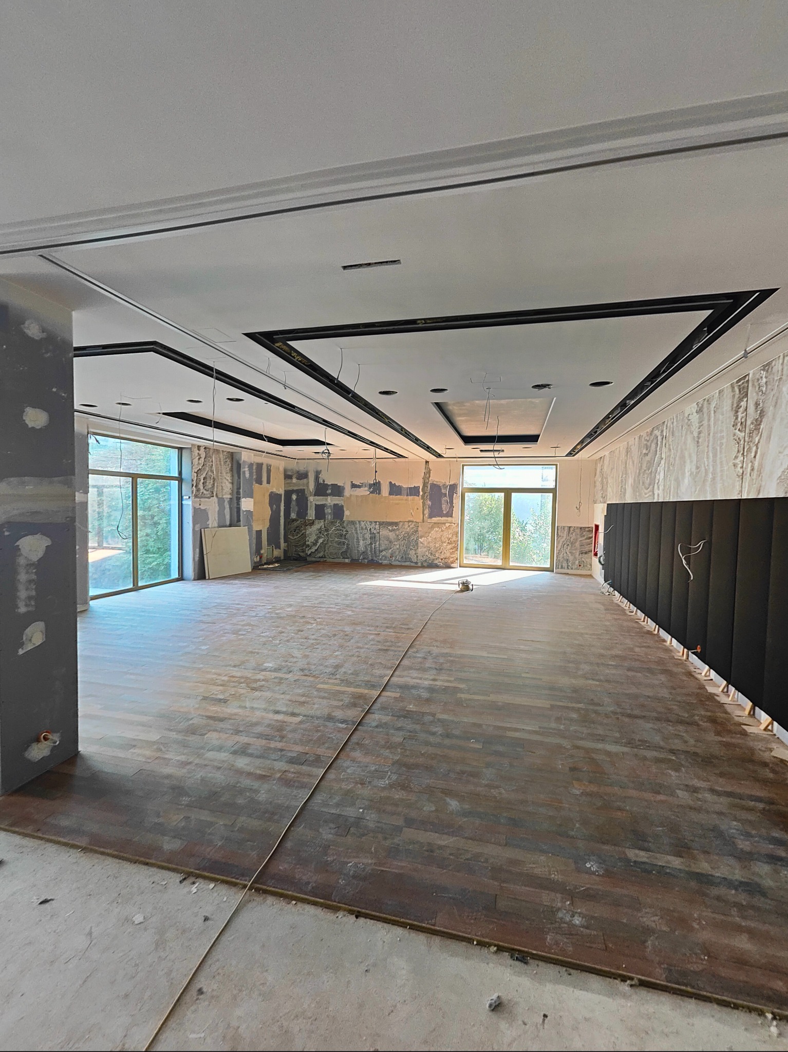 Wnętrze w trakcie prac wykończeniowych: częściowo położona drewniana podłoga, ściany przygotowane pod malowanie, widoczne okna i elementy instalacji elektrycznej pod sufitem.