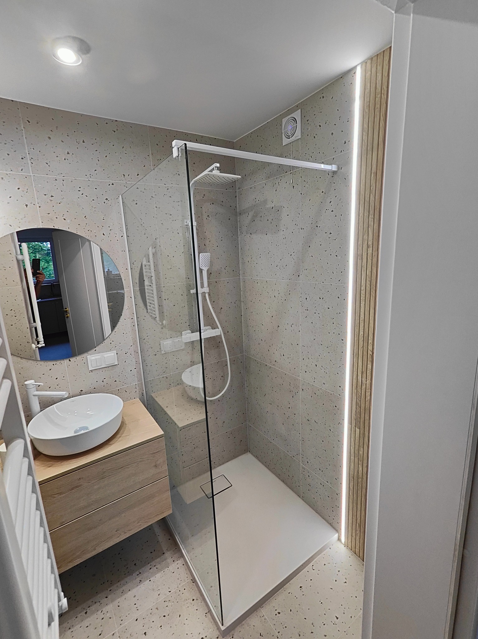 Nowoczesna łazienka po remoncie z kabiną prysznicową typu walk-in, umywalką nablatową na drewnianej szafce, okrągłym lustrem i płytkami imitującymi lastryko na ścianach i podłodze.