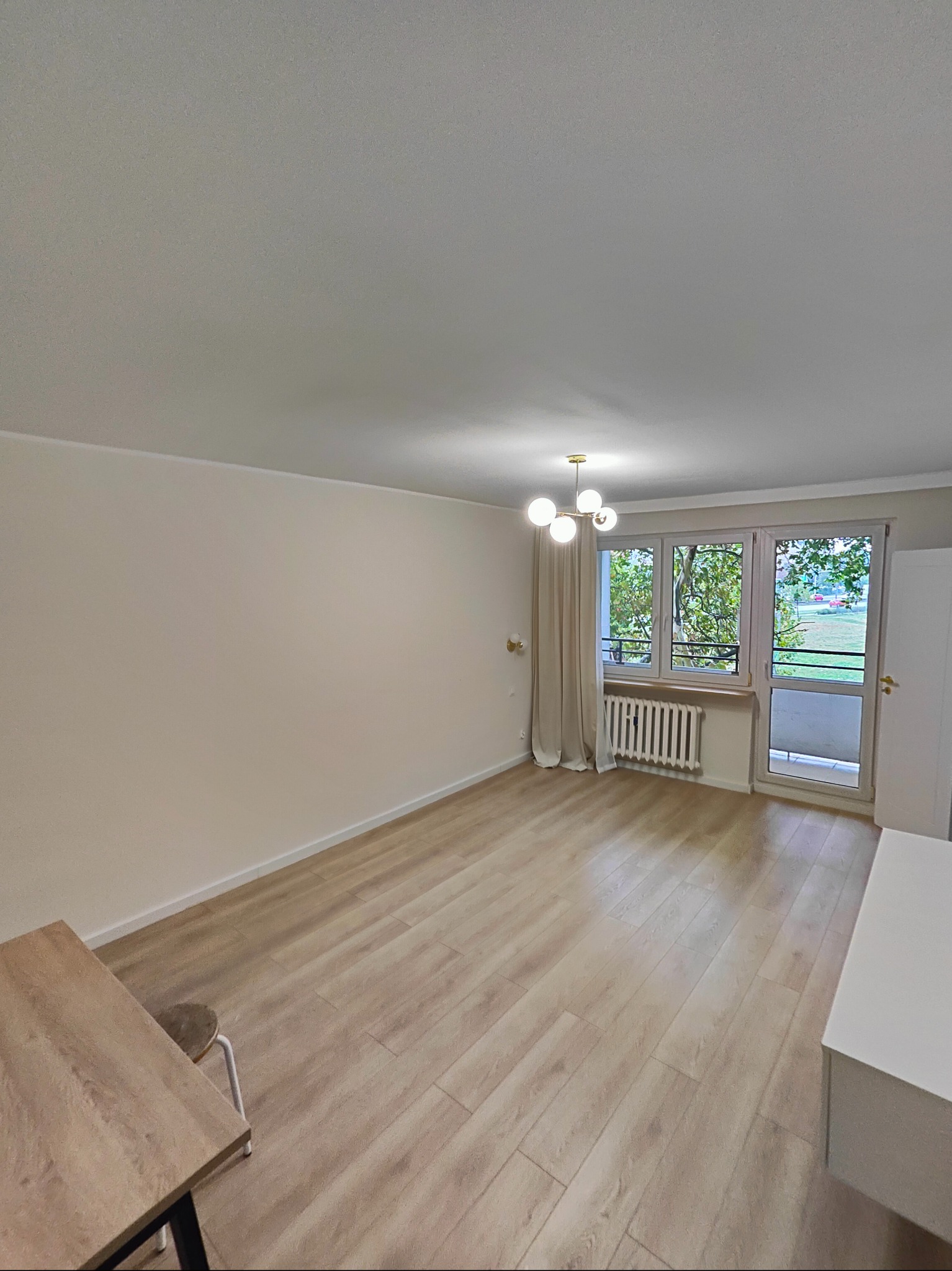 Jasne, puste pomieszczenie w Poznaniu z panelami podłogowymi, świeżo pomalowanymi ścianami w odcieniach beżu i nowoczesną lampą wiszącą. Widok na zieleń za oknem.