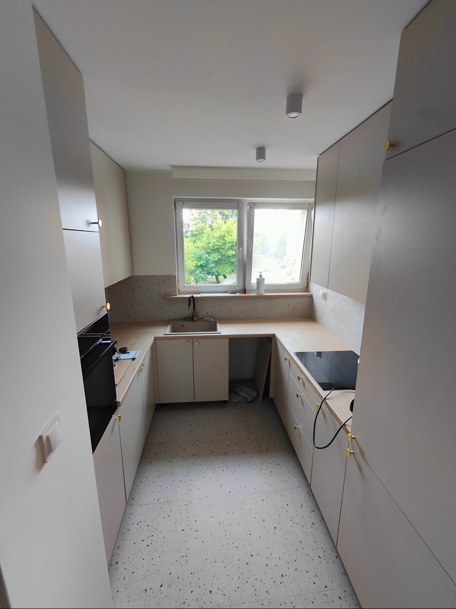 Wąska kuchnia z jasnymi, matowymi szafkami, złotymi uchwytami i blatem imitującym drewno. Uwagę zwraca podłoga w stylu lastryko oraz okno z widokiem na zieleń.