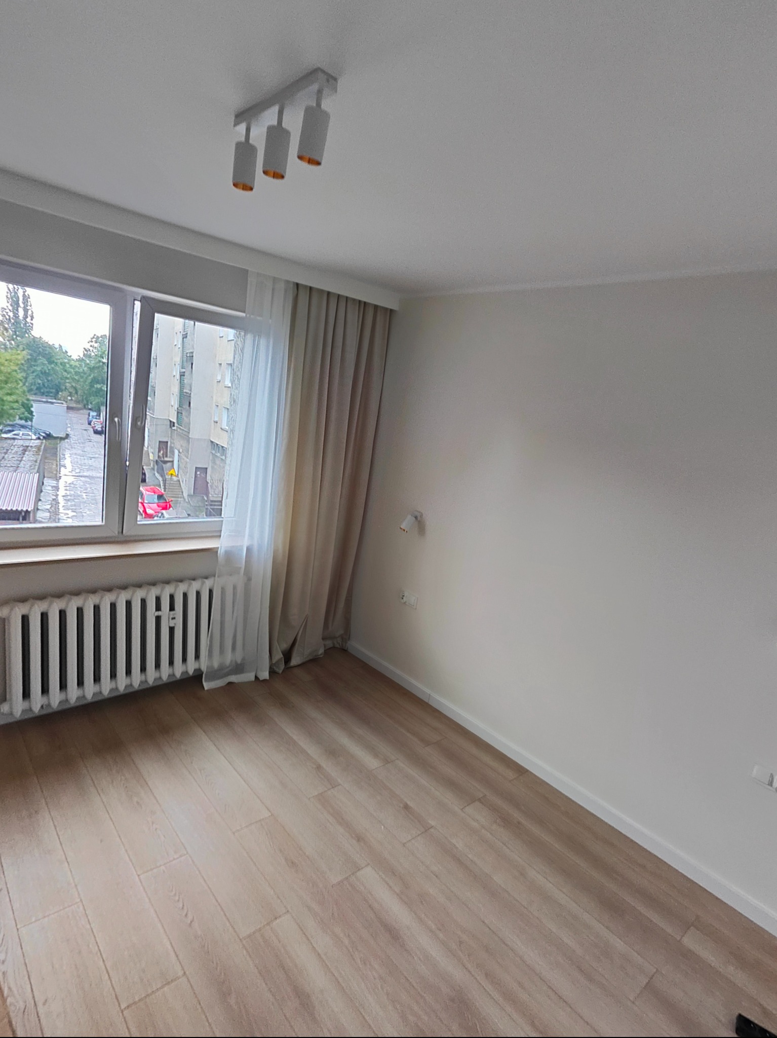 Pusty pokój po remoncie z panelami podłogowymi w odcieniach beżu, białymi ścianami i grzejnikiem pod oknem z widokiem na ulicę z czerwonym samochodem.