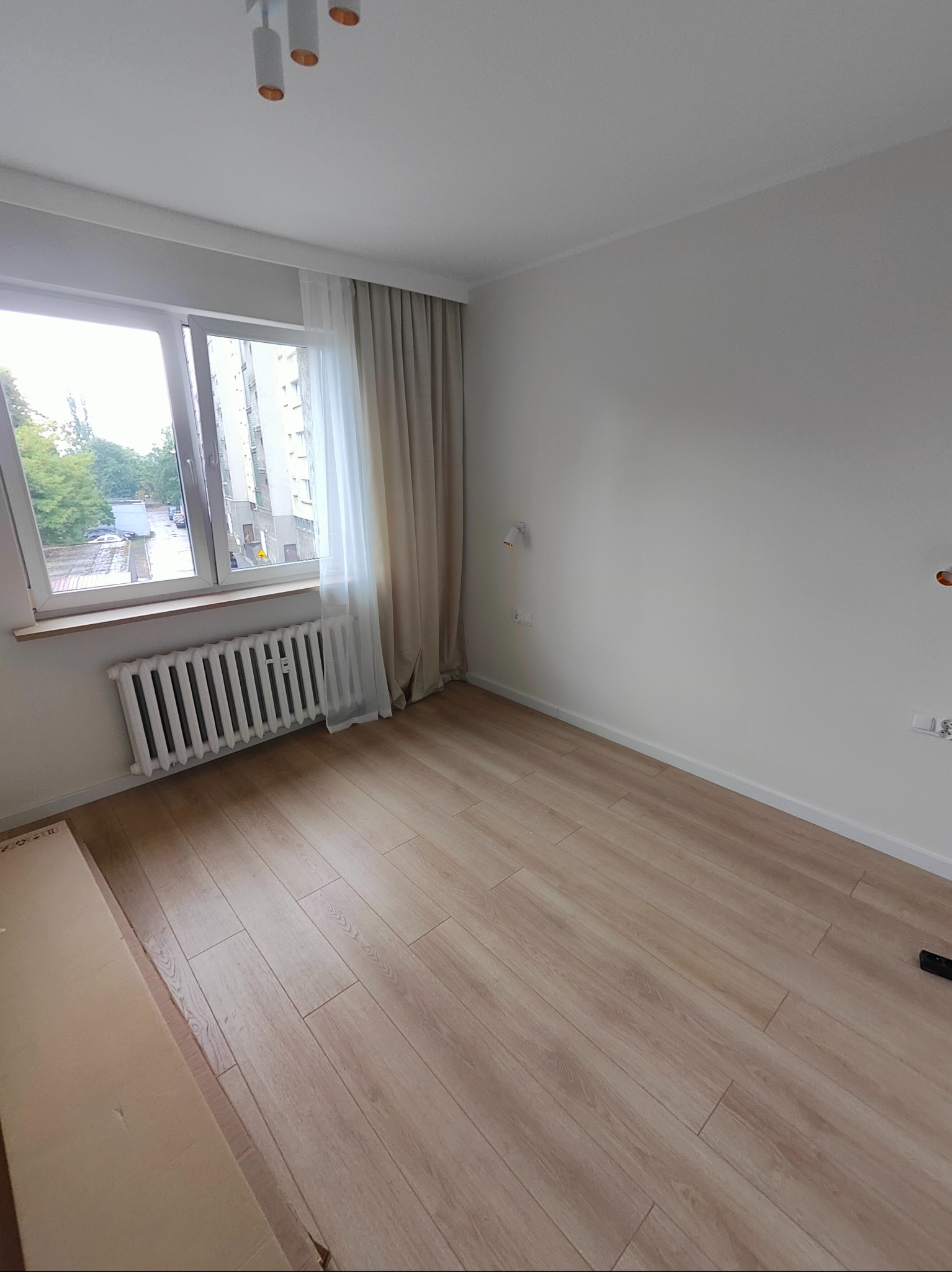 Jasne panele podłogowe ułożone w pokoju z białymi ścianami, grzejnikiem pod oknem i kartonem w rogu.