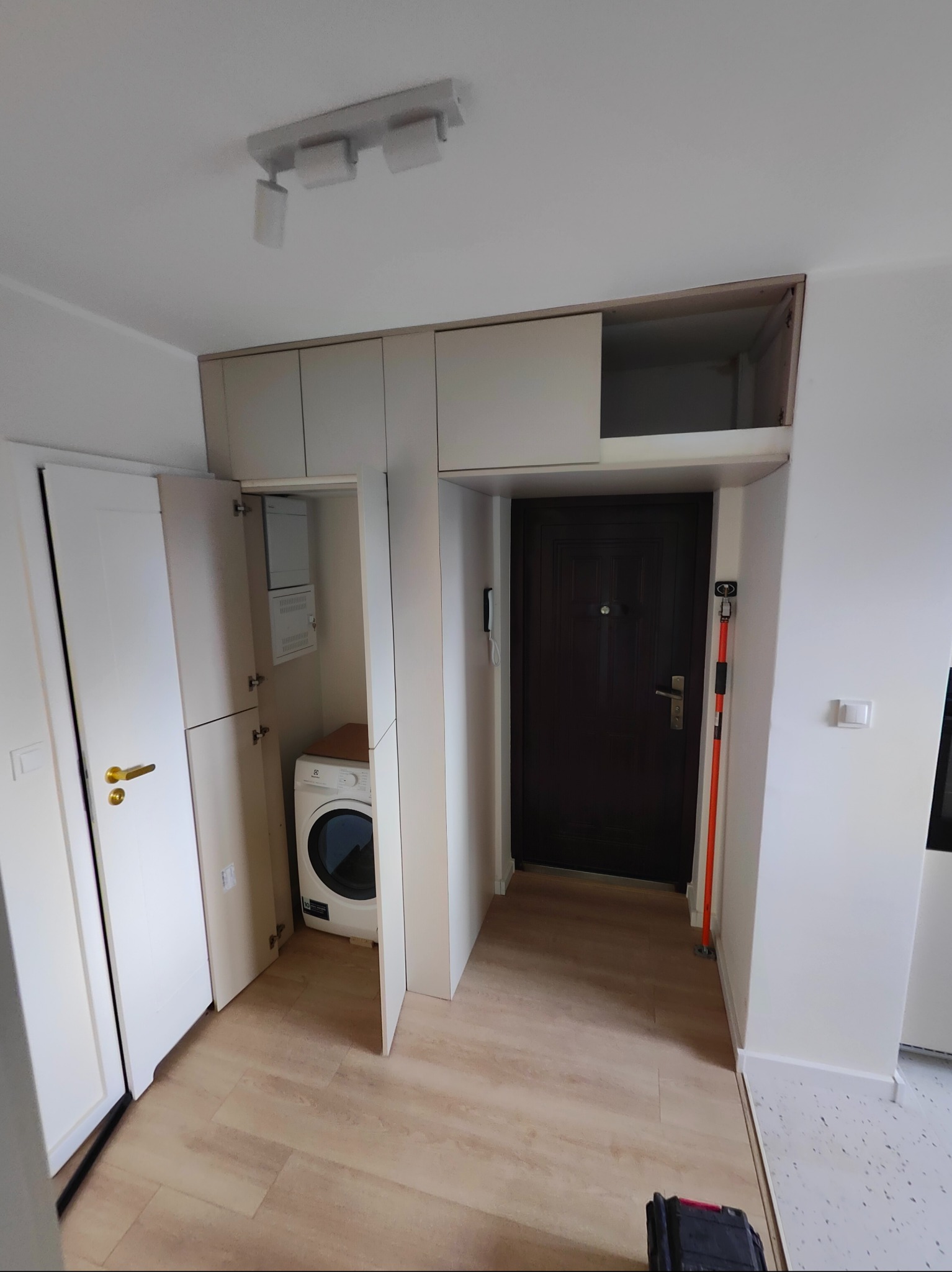 Przedpokój z zabudową meblową pod sufit, obejmującą szafki i pralkę. Widoczne panele podłogowe, drzwi wejściowe i oświetlenie sufitowe.