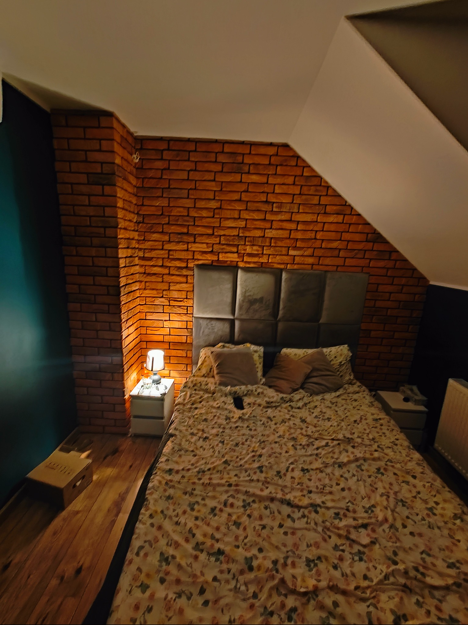 Sypialnia na poddaszu z łóżkiem, ścianą z czerwonej cegły i stolikami nocnymi z lampkami, widok z góry w słabym oświetleniu.