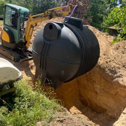 MARIUSZ WOLSKI CONSTRUCTION SERVICE - Żółto-zielona mini koparka umieszcza czarny, żebrowany zbiornik w wykopie ziemnym na tle drzew i niebieskiego nieba z chmurami.