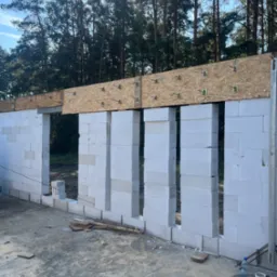 Wznoszenie ścian budynku z białych bloczków, widoczne drewniane elementy konstrukcyjne stropu, w tle las iglasty i błękitne niebo, na pierwszym planie czarne wiadro z napisem 'DAD' i butelka wody.