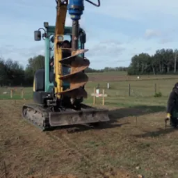 Żółto-zielona minikoparka z wiertnicą hydrauliczną, przygotowana do wiercenia otworów w ziemi na działce z wyznaczonymi punktami, widoczny operator i pracownik w oddali.