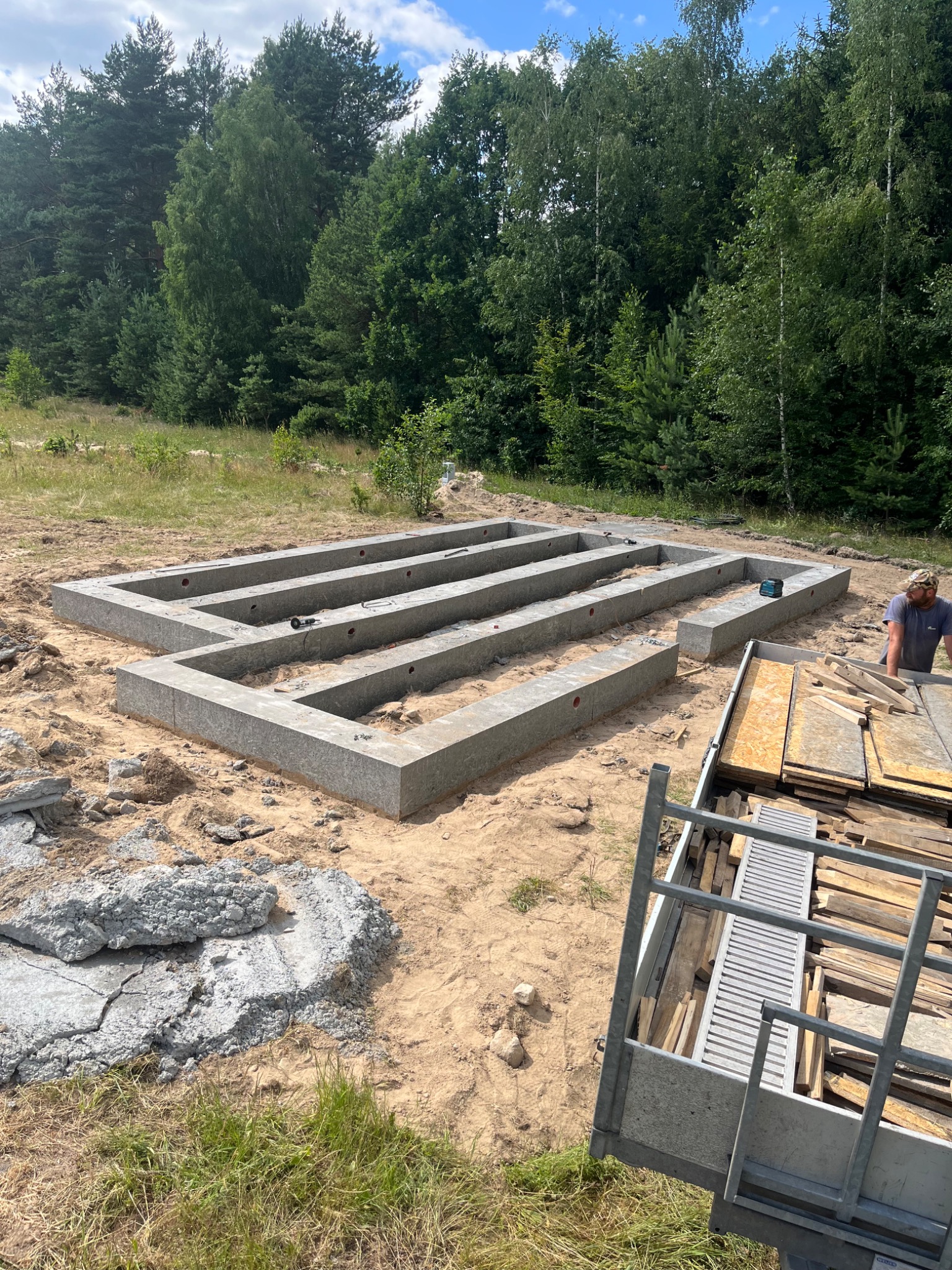 Szary fundament w formie prostokąta z wewnętrznymi belkami, widoczne otwory na instalacje, teren piaszczysty z resztkami gruzu, w tle las.